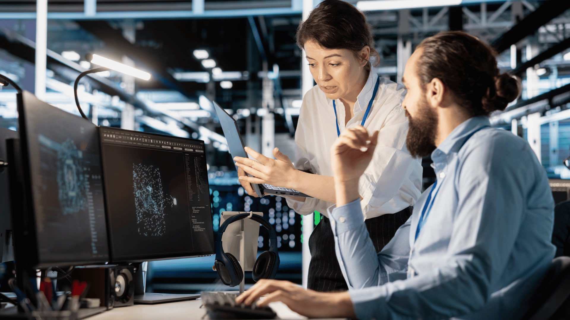 Two people collaborating in a modern tech workspace with dual monitors displaying digital graphics and a laptop.