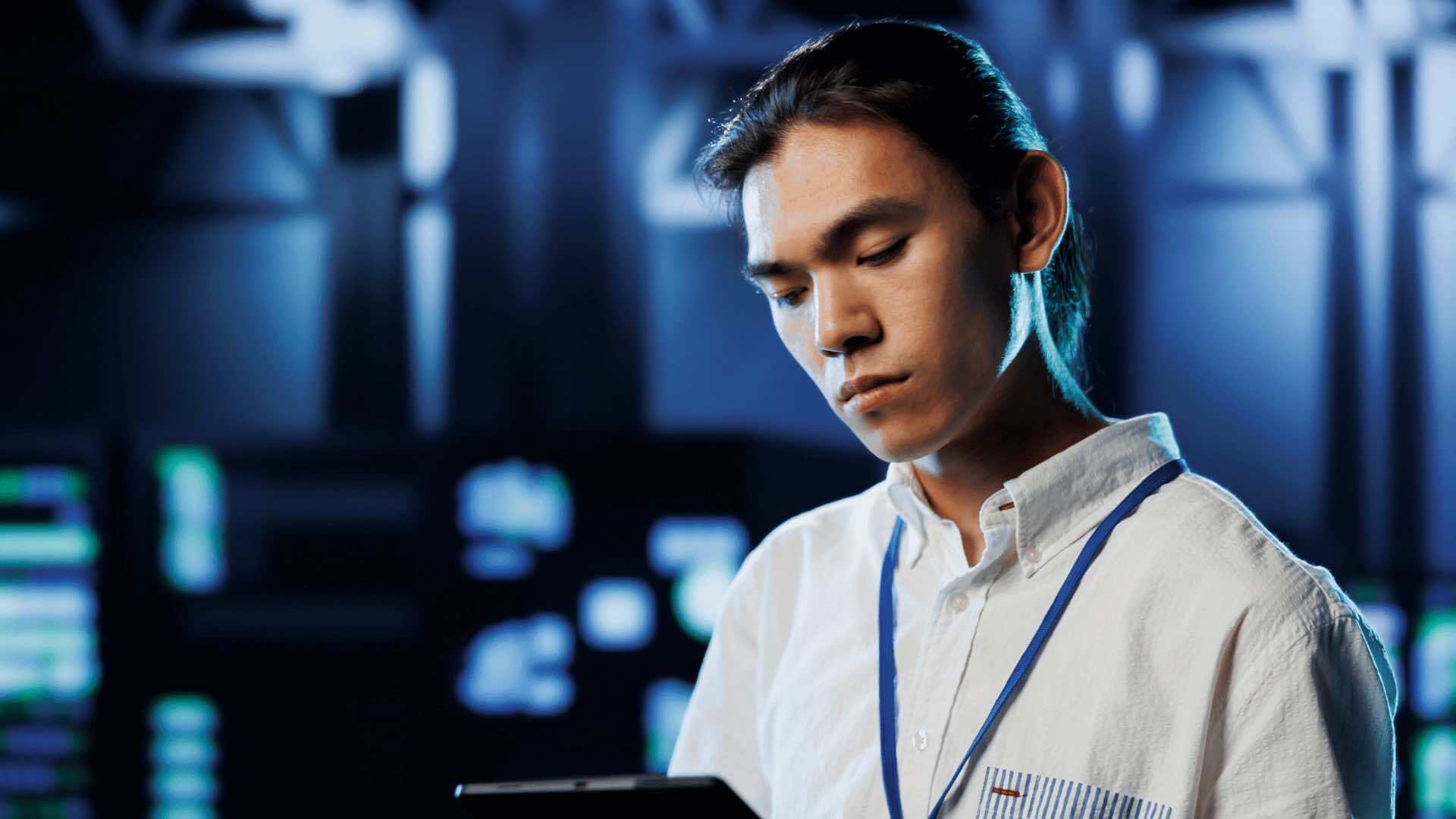 An IT expert wearing a white collared shirt with a lanyard, holding a tablet in a data center.