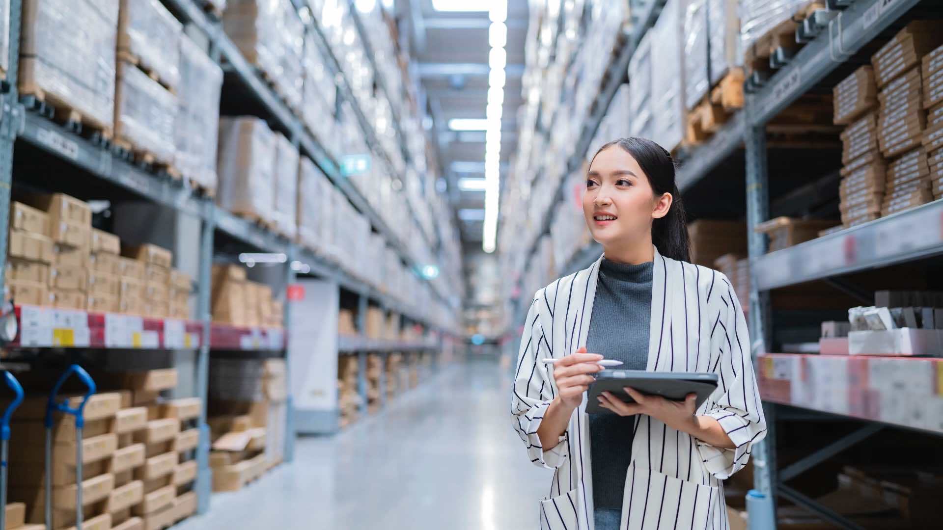 A person holding a tablet in a large warehouse aisle filled with shelves stacked with boxes and packages, indicating an inventory or logistics setting.
