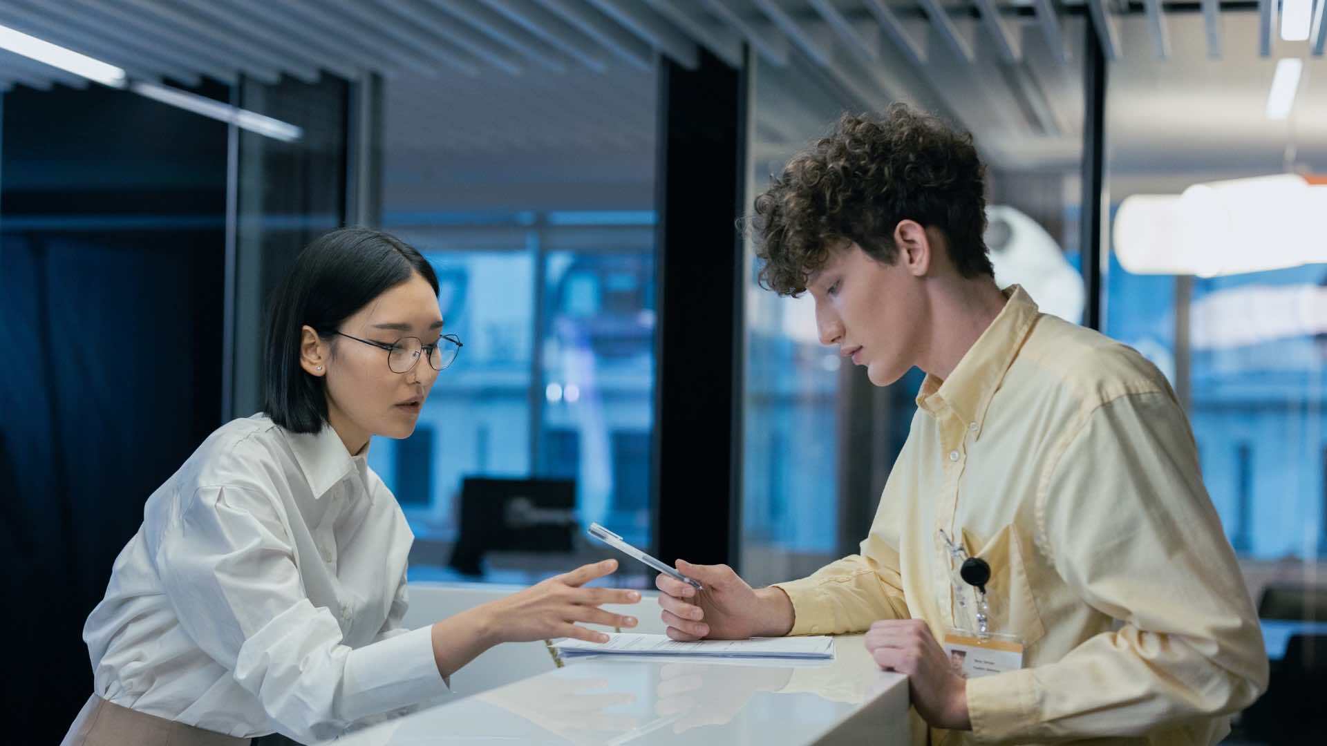 Hai người trong không gian văn phòng hiện đại, đang trao đổi tại quầy. Một người cầm điện thoại thông minh trong khi người kia ra hiệu, gợi lên cuộc trò chuyện hợp tác.