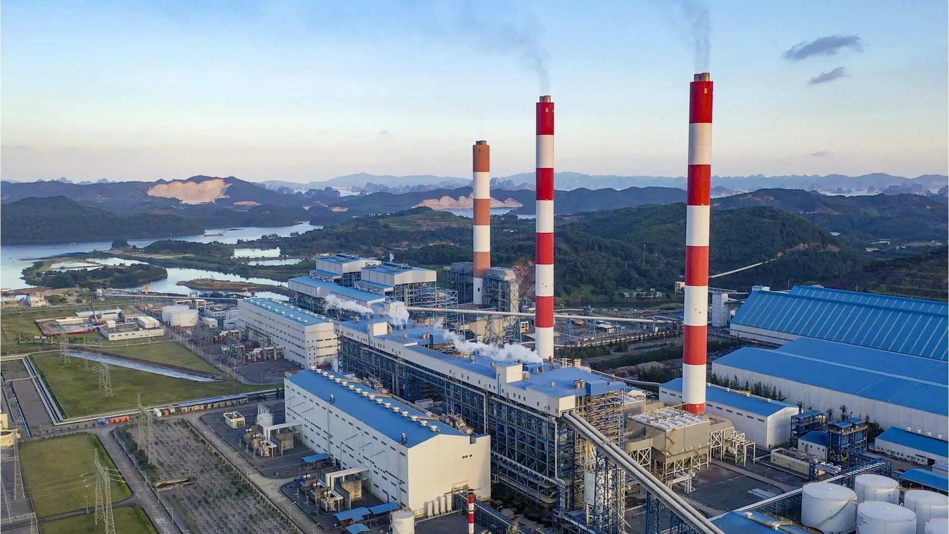 Một nhà máy công nghiệp lớn với các ống khói đỏ trắng thải khói, xung quanh là tòa nhà và bồn chứa, phía sau là núi và mặt nước.